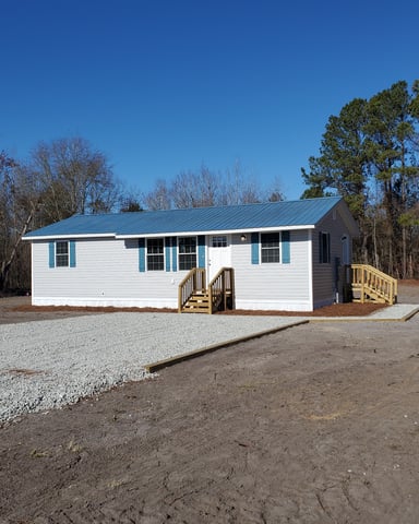 White single-wide mobile home with blue metal roof and turquoise shutters, wooden deck and steps, surrounded by bare and evergreen trees.