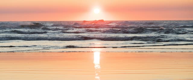 Warm peach and gold sunset reflected on calm Atlantic Ocean waves lapping a flat sandy beach